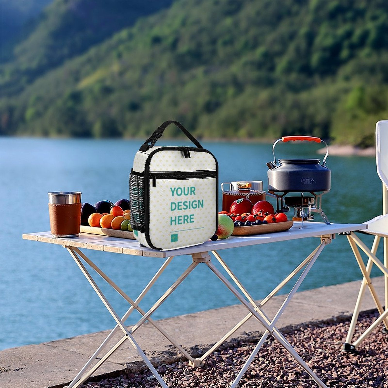 Customizable lunch tote bag with 'YOUR DESIGN HERE' on a picnic table by the lake, perfect for work or travel.