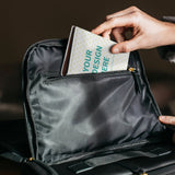 Custom passport holder with 'YOUR DESIGN HERE' text, placed in a black bag. PU material travel gift.