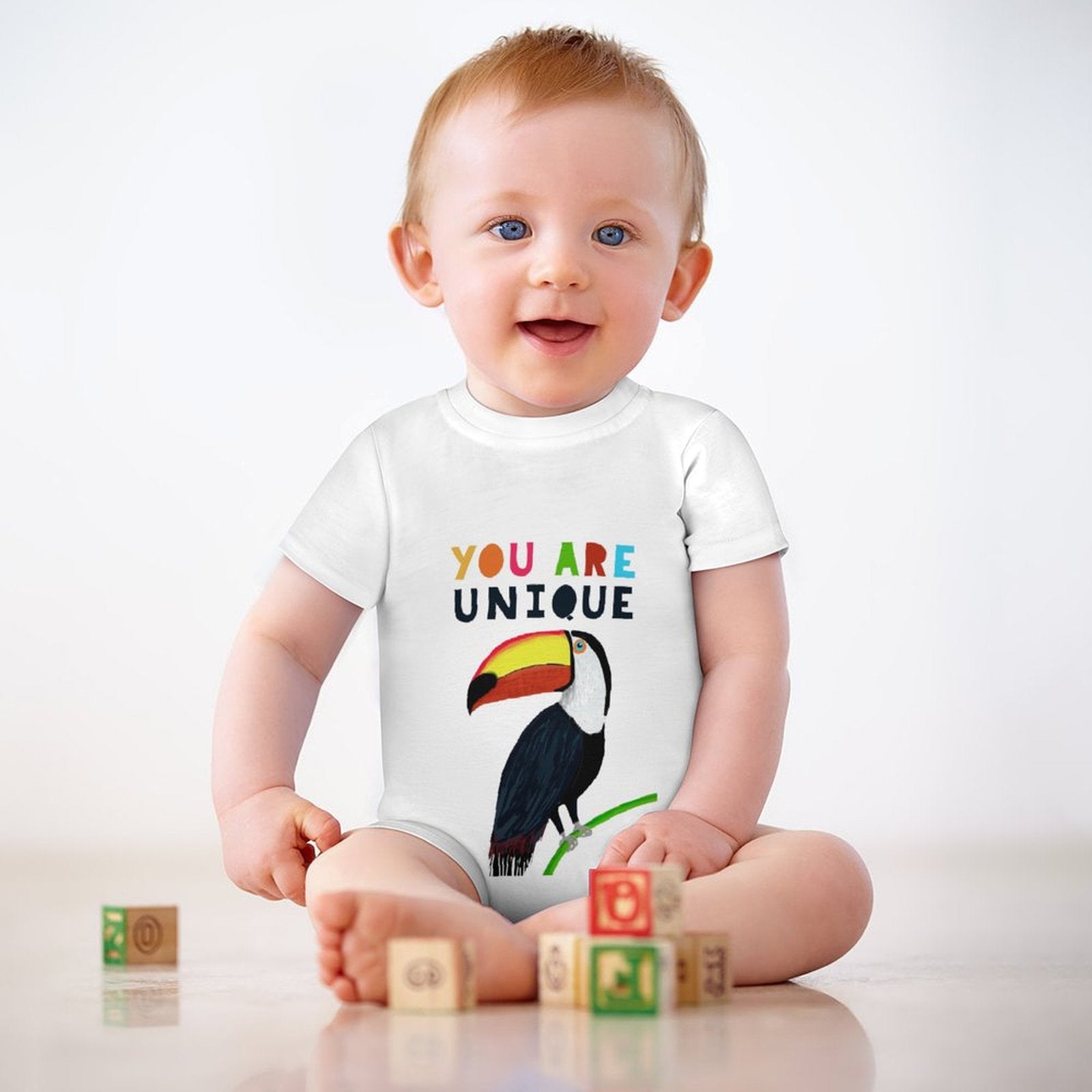 Baby in soft cotton bodysuit with toucan graphic and 'YOU ARE UNIQUE' text. Perfect baby gift 0-24 months.