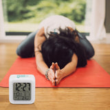 Customizable white digital alarm clock, time, date, temp display, beside a woman meditating on a yoga mat.