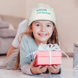 Customizable baby beanie hat with 'YOUR DESIGN' text, held by happy girl with gift, perfect for kids.