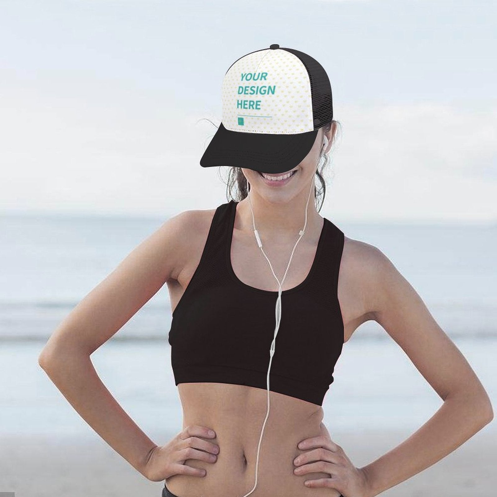 Customizable trucker hat with 'YOUR DESIGN HERE' text, black bill, and mesh back on a woman by the beach.