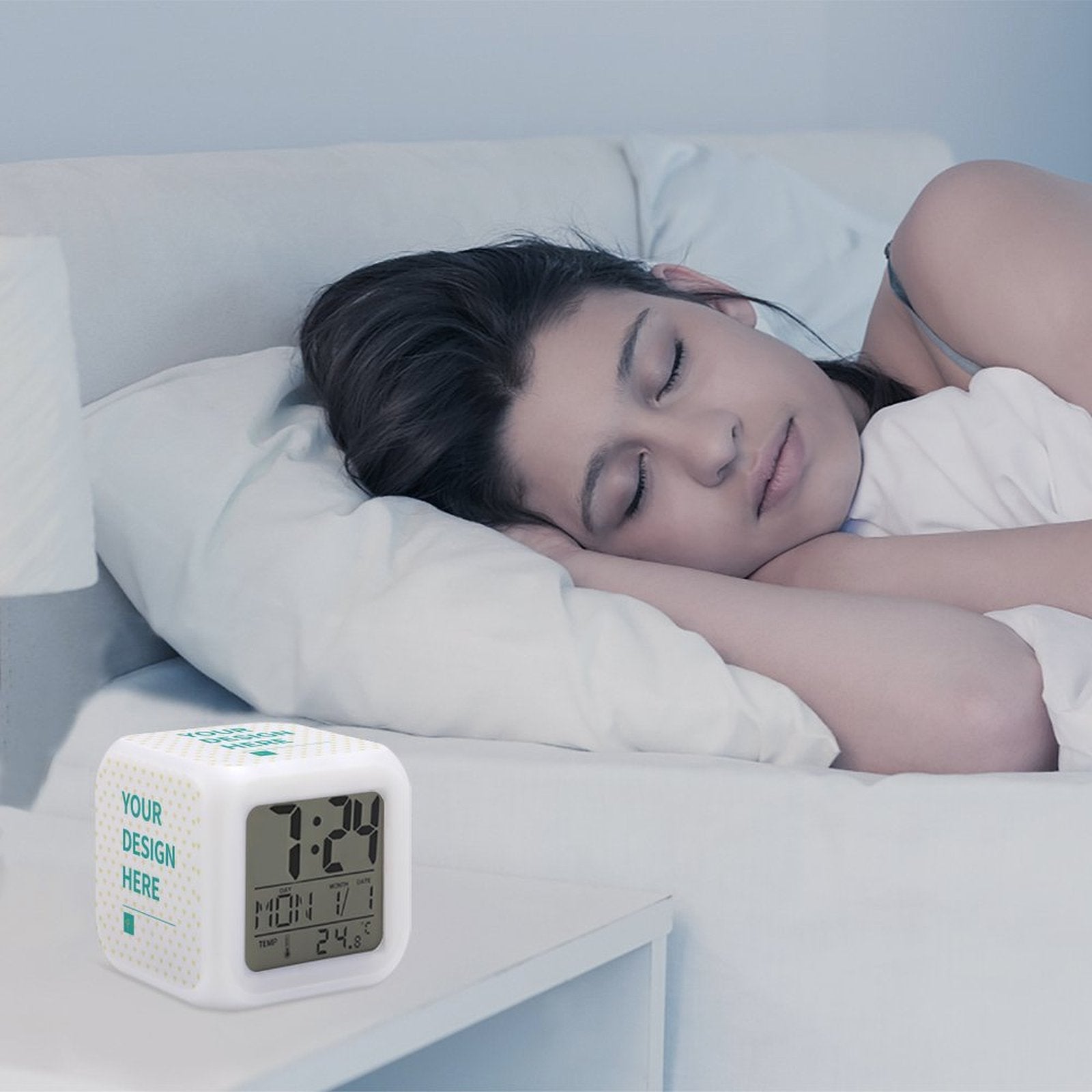 Customizable digital alarm clock beside a sleeping woman. White cube alarm with time, date, and design display.