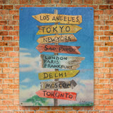 Metal sign with international destinations including Los Angeles, Tokyo, and more, displayed against a brick wall.