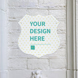 Customizable shield-shaped metal sign with 'YOUR DESIGN HERE' on white brick wall, perfect for shop decor.